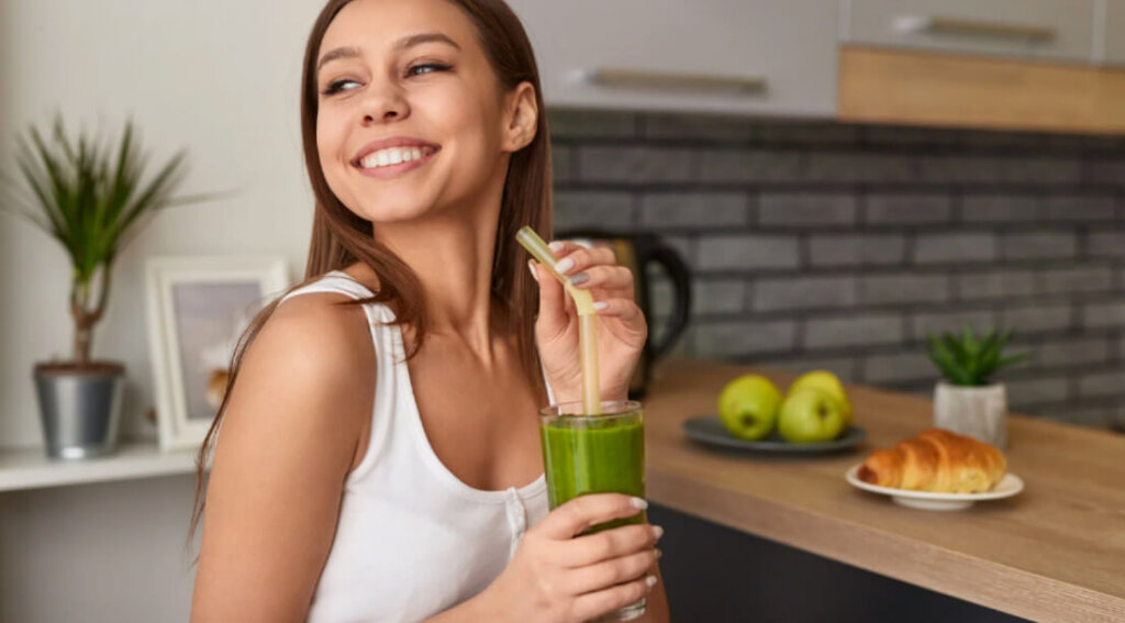 A woman smiles while holding a green smoothie in a bright kitchen, representing healthy lifestyle changes during recovery.