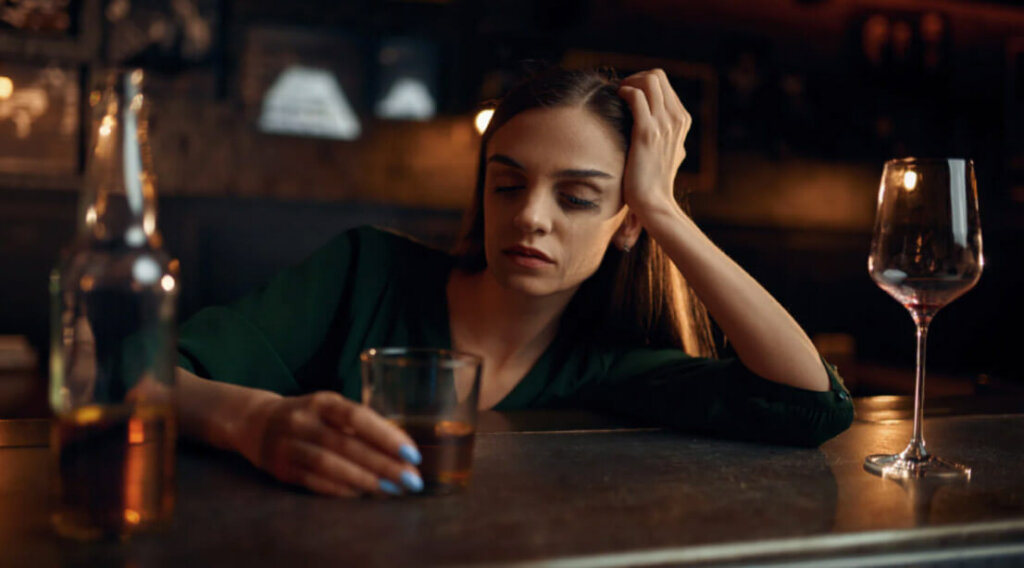 A woman sits alone at a bar looking distressed while holding a drink, illustrating the challenges of alcohol dependence.