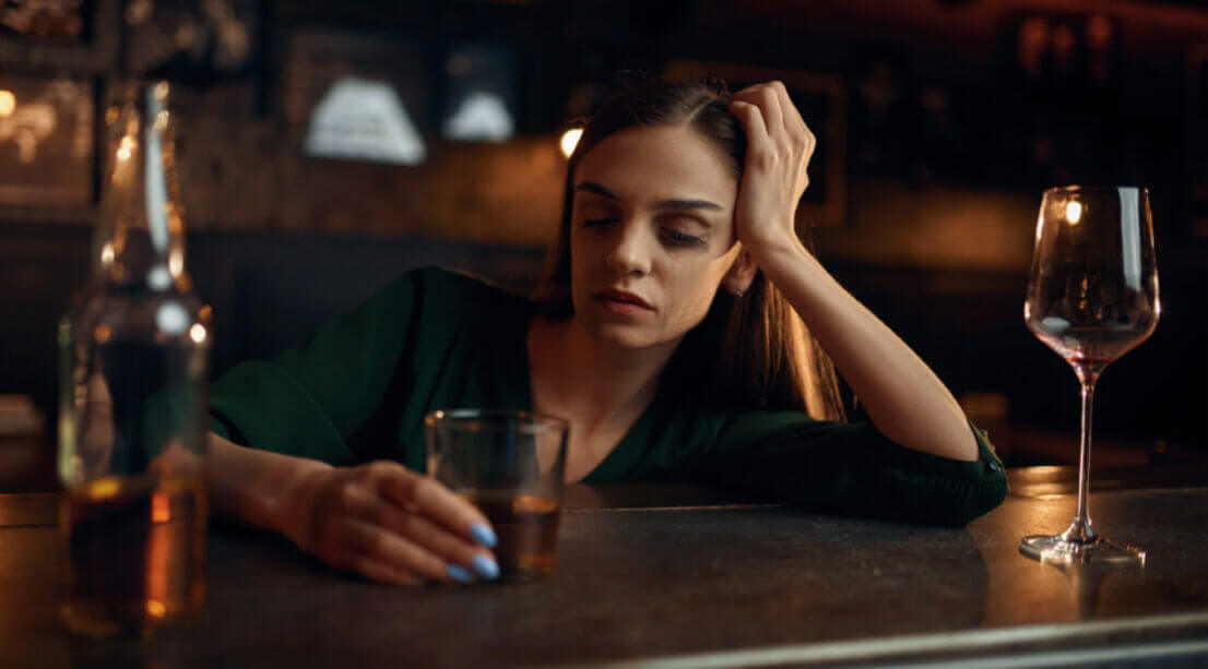 A woman sits alone at a bar looking distressed while holding a drink, illustrating the challenges of alcohol dependence.