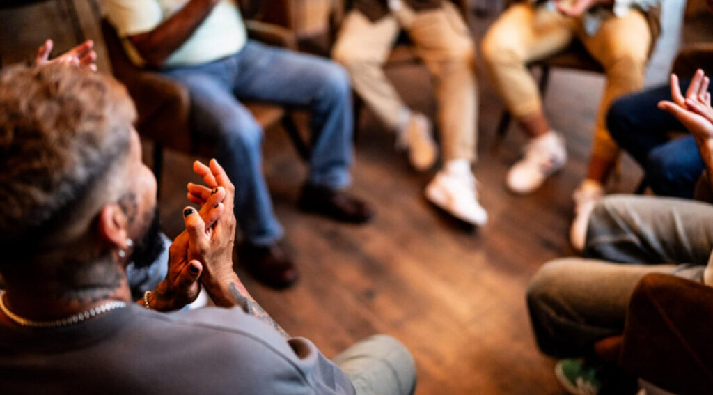 A group of people sit in a circle during a support meeting, actively engaging in discussion and shared experiences.