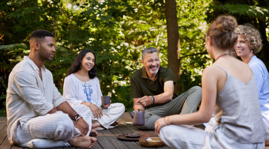 A diverse group of adults sit together outdoors in a circle, sharing conversation and support in a peaceful setting.