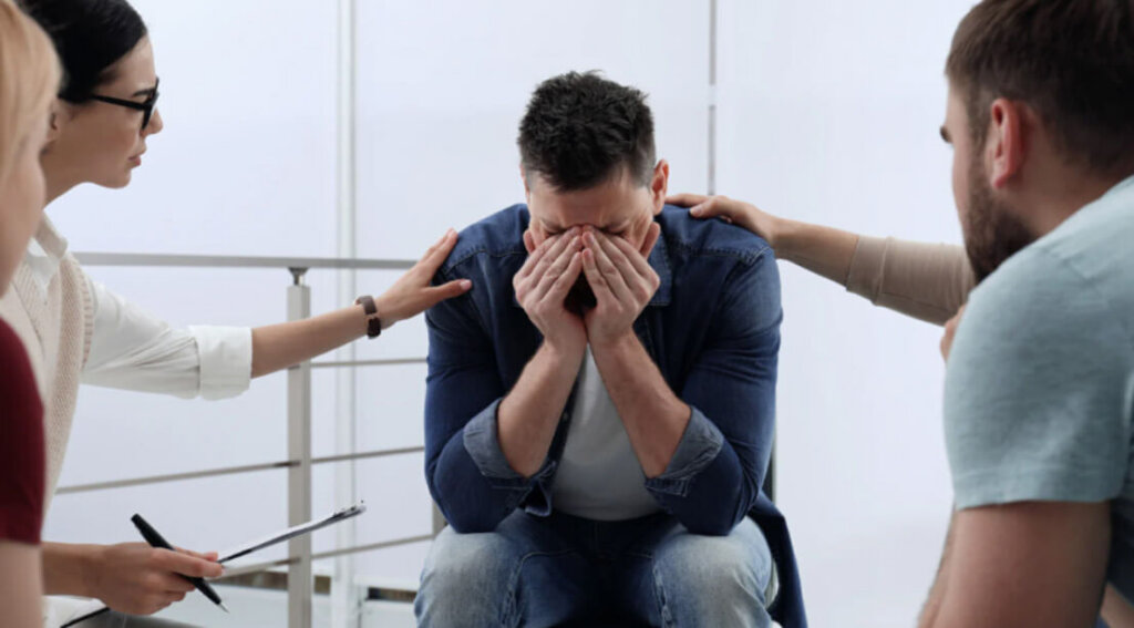 A man sits with his face in his hands while others offer support, showing emotional struggle and compassionate care.