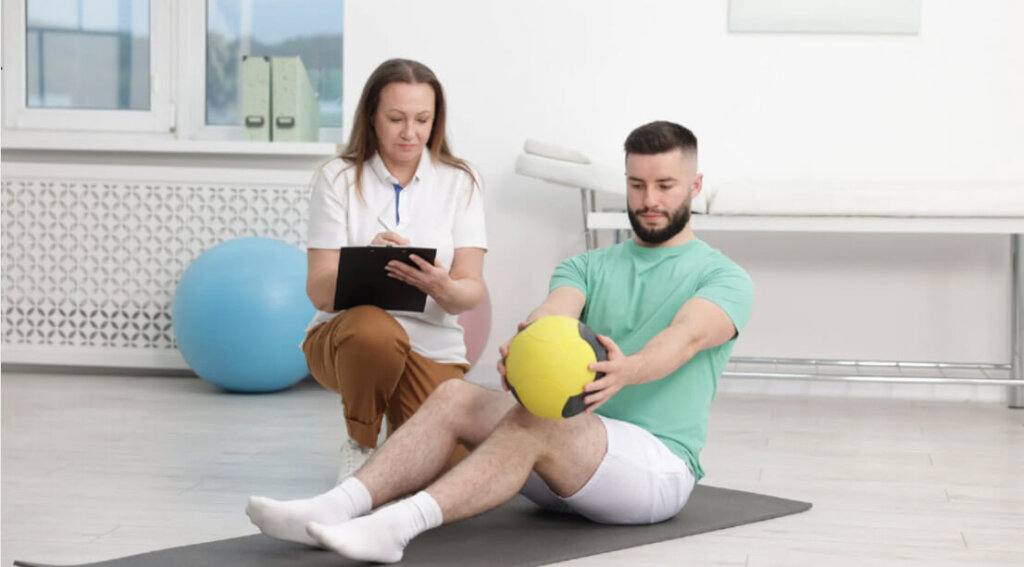 A therapist observes and takes notes while a man performs a seated exercise with a medicine ball during a supervised session.