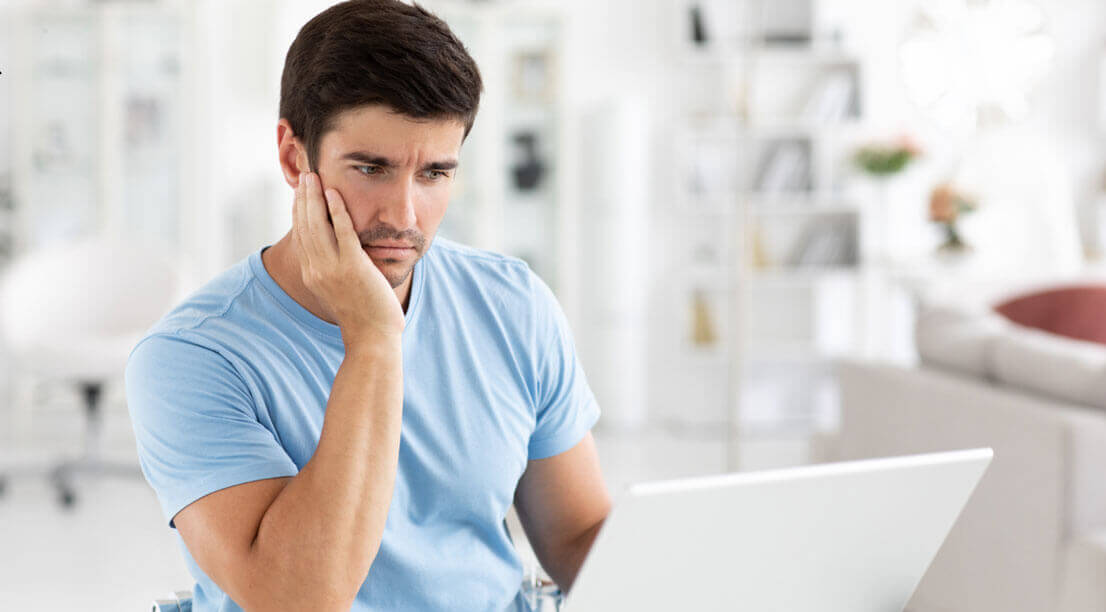 A man sitting at a laptop looking worried and deep in thought while researching options.