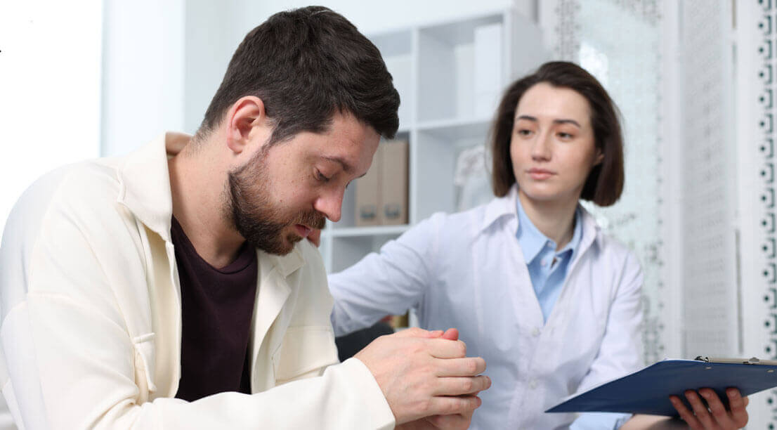A counselor offering support to a man who looks down, appearing thoughtful and emotional during therapy.