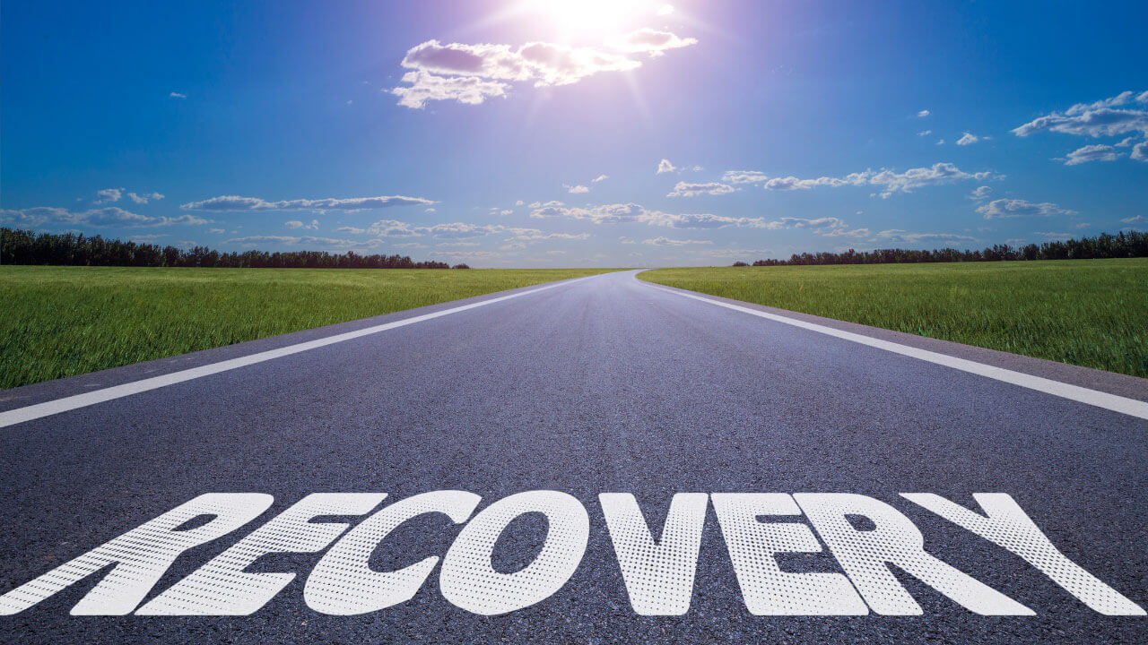 An open road stretching toward the horizon under a bright sun and blue sky, with the word 'RECOVERY' painted in large white letters across the pavement.