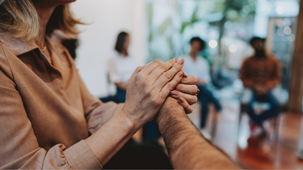 A close-up of two people clasping hands warmly during a group therapy session, with several blurred participants seated in a circle in the background.
