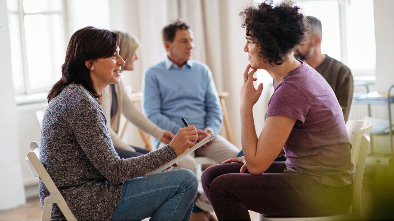 A woman with a clipboard listens attentively to a young person with curly hair during a group therapy session, with other participants seated nearby in a bright room.