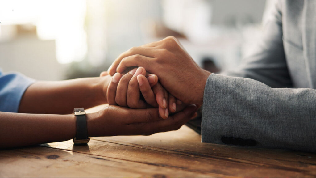 A close-up of two people's clasped hands resting on a wooden table, one wearing a smartwatch and the other in a gray blazer, conveying comfort and support.