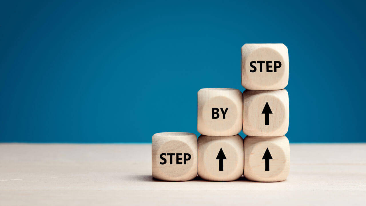 Wooden blocks stacked like ascending steps spell out 'STEP BY STEP' with upward arrows, set against a blue background.