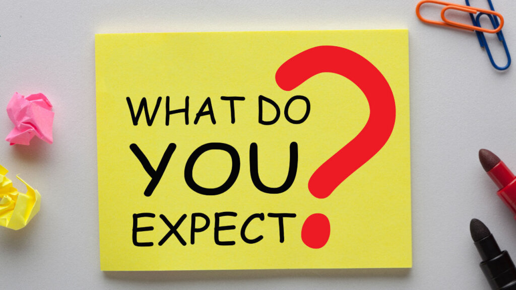 A yellow sticky note reading 'WHAT DO YOU EXPECT?' in bold black letters with a red question mark, surrounded by crumpled colorful paper and office supplies.