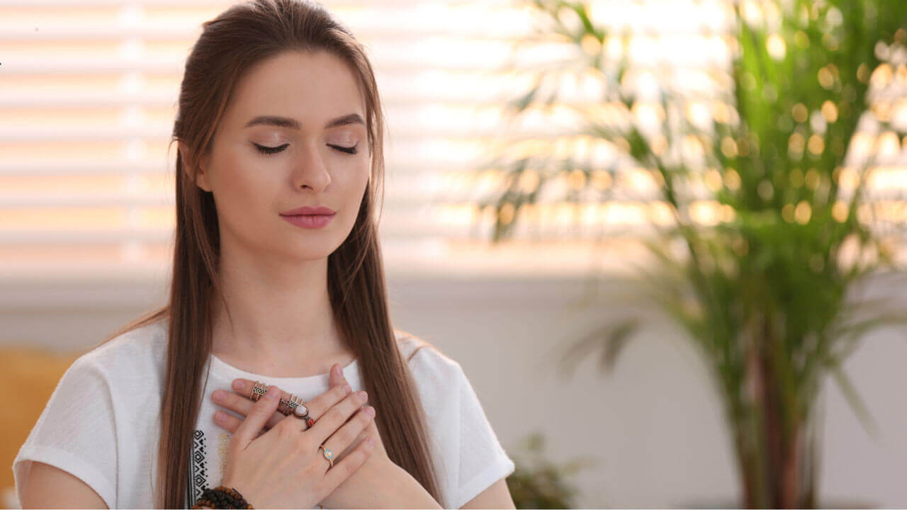 A woman with long brown hair and closed eyes rests her clasped hands over her heart in a calm, meditative pose, with soft window light and a potted plant in the background.