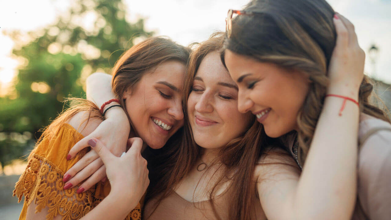 Three friends embrace and smile together outdoors in a warm display of support.