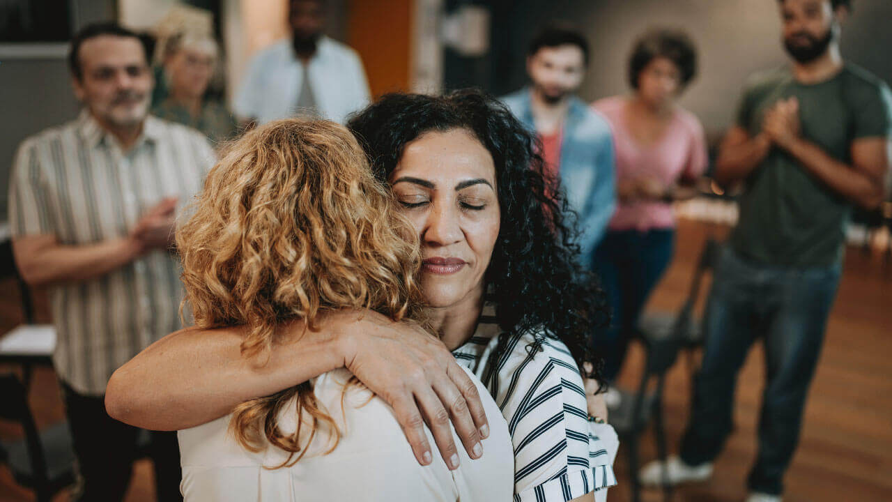 Two women embrace as members of a recovery support group applaud in the background.