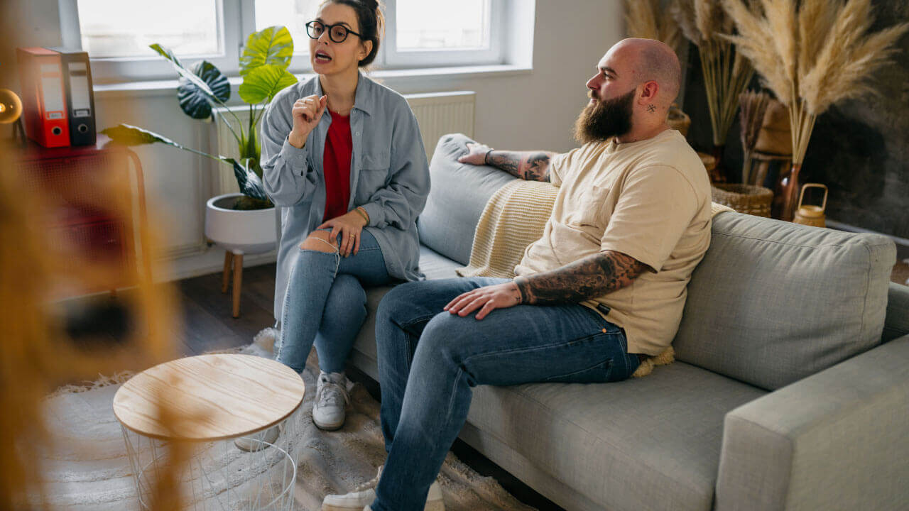 Woman gesturing while speaking to a bearded tattooed man seated together on a couch.