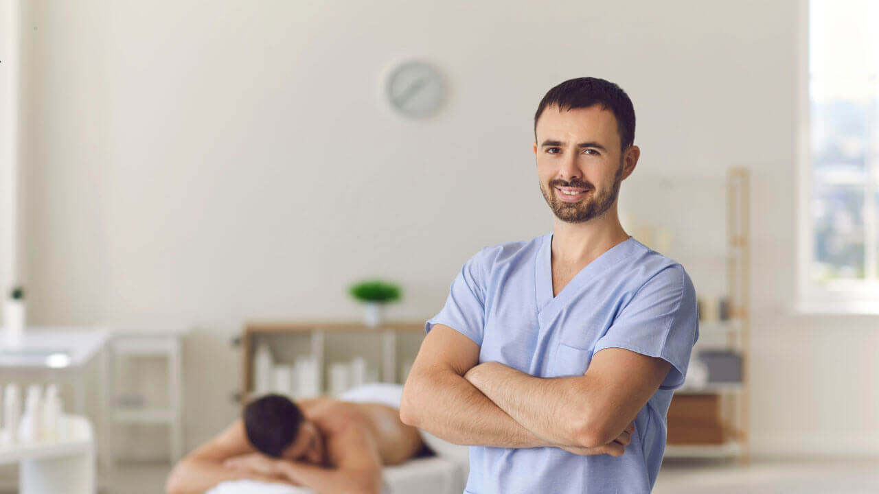 A healthcare professional stands confidently in a bright treatment room with a patient resting behind him.