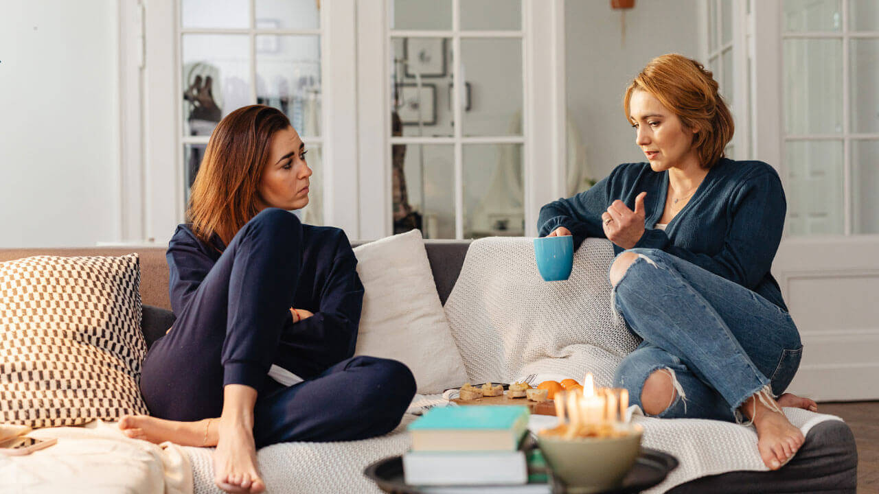 Two women sit on a couch having a serious conversation with tea and candles on the table.