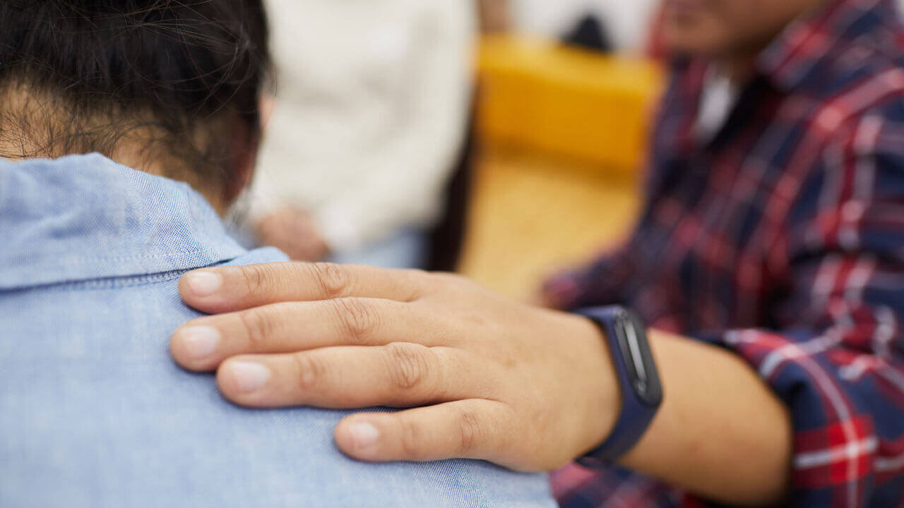 Close up of a hand resting gently on someone's shoulder from behind during a group setting.