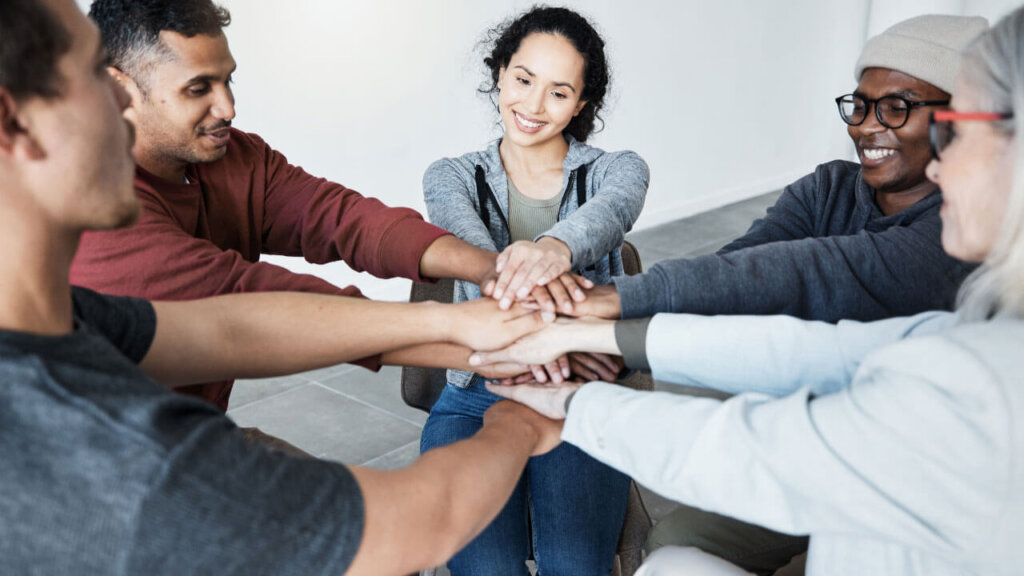 A diverse group of people stack their hands together in a show of support and unity during a group therapy session.