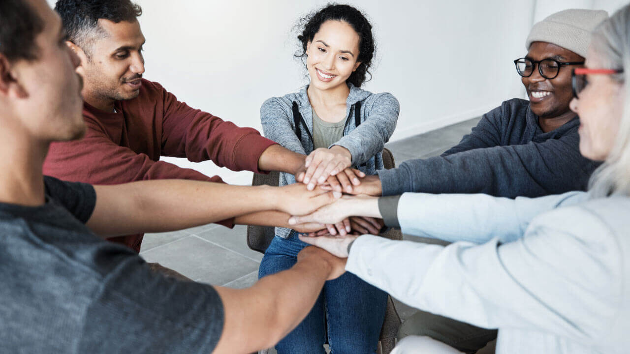 A diverse group of people stack their hands together in a show of support and unity during a group therapy session.