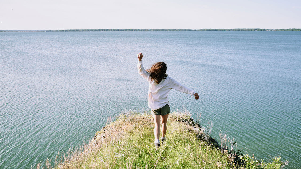 A woman with her arms outstretched stands on a grassy cliff overlooking a wide, calm lake.