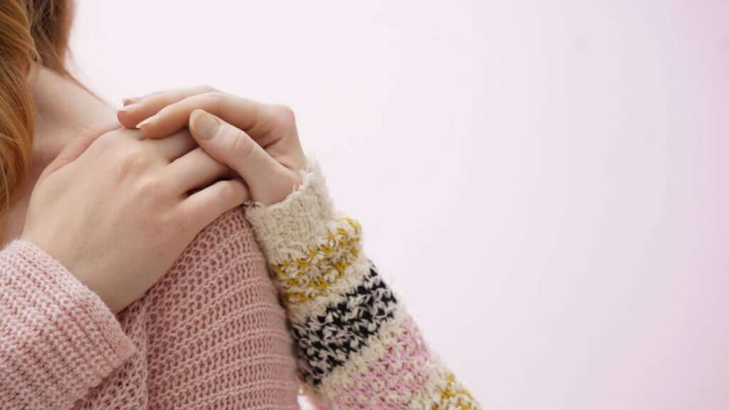 A close-up of a woman in a pink knit sweater clasping her hands against her shoulder.