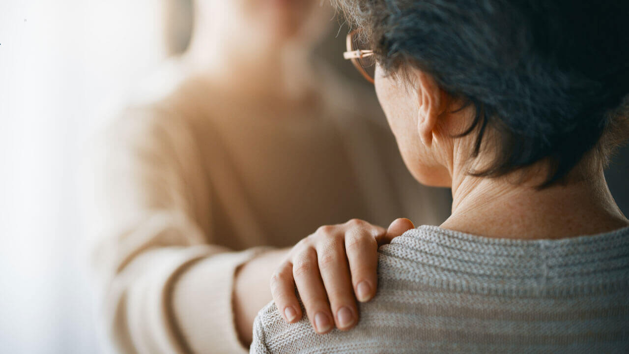 A person rests a comforting hand on another person's shoulder in a softly lit room.