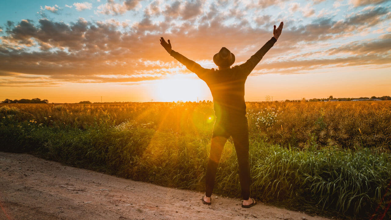 A person in a hat raises both arms toward a glowing sunset over a golden field.