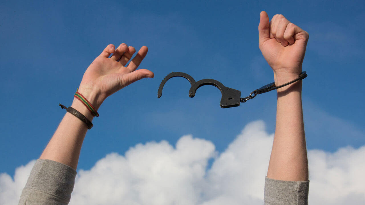 Two raised arms break free from open handcuffs against a bright blue sky, symbolizing freedom from addiction.