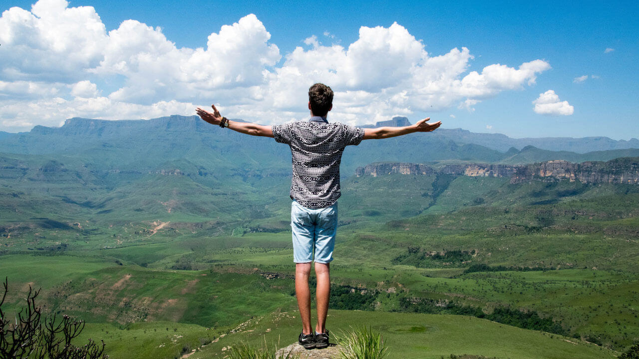 A man stands with arms spread wide on a rock overlooking green mountains and a blue sky.