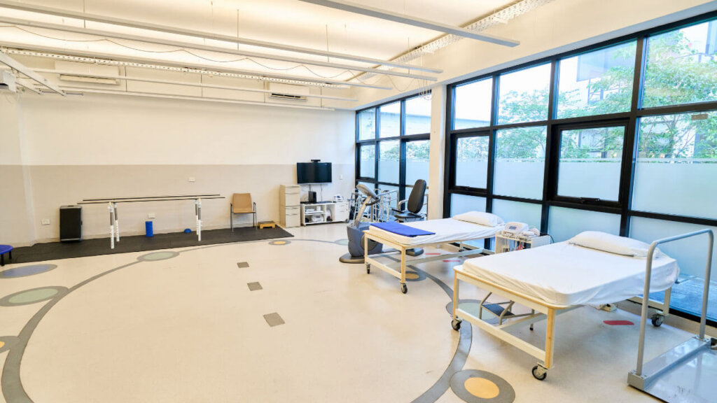 A bright rehabilitation room with two treatment beds, parallel bars, and large windows.