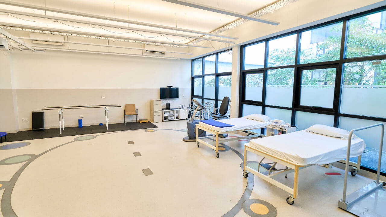 A bright rehabilitation room with two treatment beds, parallel bars, and large windows.